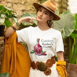 Può includere: Una donna indossa una maglietta bianca con una grafica di una bottiglia di cordiale ai lamponi e la scritta "Cuthbert Raspberry Cordial". Indossa un cappello marrone, orecchini d'oro e una cintura marrone larga con dettagli metallici.