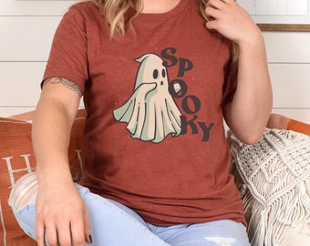 May include: A rust-colored t-shirt featuring a cartoon ghost with the word "Spooky" in a retro font. The ghost is light green and white. The shirt is worn by a person.