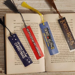 May include: Four colorful bookmarks with tassels rest on an open book. The bookmarks feature the text "A Charlie Brown Christmas", "Spider-Man", "Home Alone 2", and "Lord of the Rings".