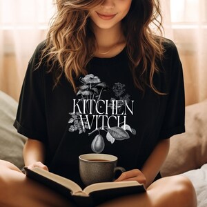 May include: A black t-shirt with a white graphic design of a pear, mushrooms, and leaves. The text "Kitchen Witch" is printed in white on the shirt.