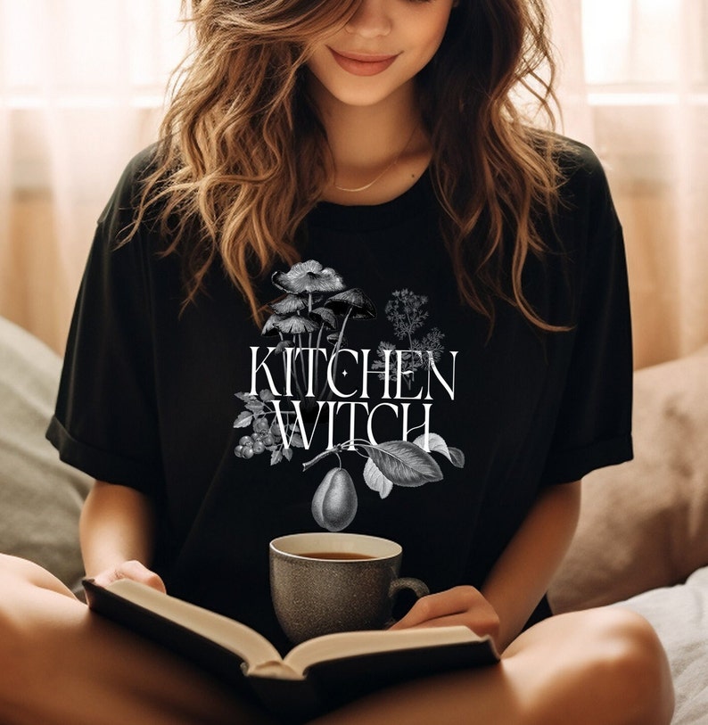 May include: A black t-shirt with a white graphic design of a pear, mushrooms, and leaves. The text "Kitchen Witch" is printed in white on the shirt.