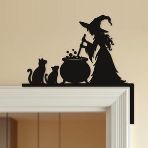 May include: A black metal door corner topper featuring a silhouette of a witch wearing a pointed hat, standing beside a bubbling cauldron and stirring it with a long spoon, accompanied by two cats sitting side by side on a flat horizontal base.