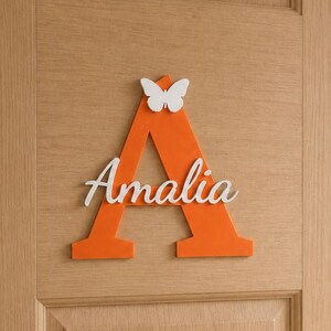 May include: A large orange wooden letter A mounted on a light wood door. The letter features a white butterfly cutout at the top and the name Amalia in white cursive script lettering across the center of the letter. The sign is flat and matte.