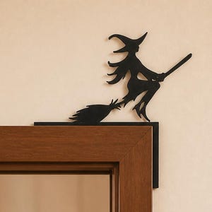 Puede incluir: Una silueta de metal negro de una bruja montada en una escoba, diseñada para montarse en la esquina del marco de una puerta. La figura plana y recortada muestra un sombrero puntiagudo, cabello largo y una escoba sobre el marco de madera.