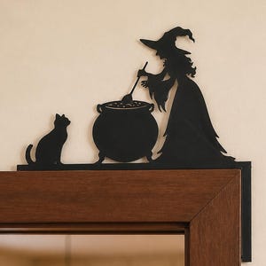 Puede incluir: Un adorno de metal negro para marco de puerta con la silueta de una bruja con sombrero puntiagudo, revolviendo un caldero grande con una cuchara, y un gato sentado junto a la olla. La pieza decorativa está diseñada para la esquina del marco.