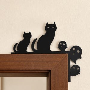 Puede incluir: Una decoración de esquina de puerta de metal negro con la silueta de dos gatos y tres fantasmas. El diseño está formado para encajar sobre la esquina superior del marco de una puerta, con los gatos en el borde superior y los fantasmas al lado.