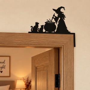 May include: A black metal door corner sign featuring a silhouette of a witch wearing a pointed hat, standing beside a bubbling cauldron and two cats. The decorative piece is designed to be mounted on the upper corner of a door frame.