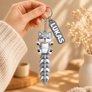 May include: A gray and white wolf keychain featuring a cartoon wolf character with a long, segmented tail and a rectangular tag with the name LUKAS in white raised letters, all attached to a silver metal split ring and a short chain.