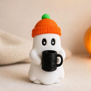 May include: A white ghost figurine wearing an orange knit beanie with a green pom-pom. The ghost has large black eyes and is holding a small black mug decorated with embossed spiderweb and bat patterns in its two rounded arms.