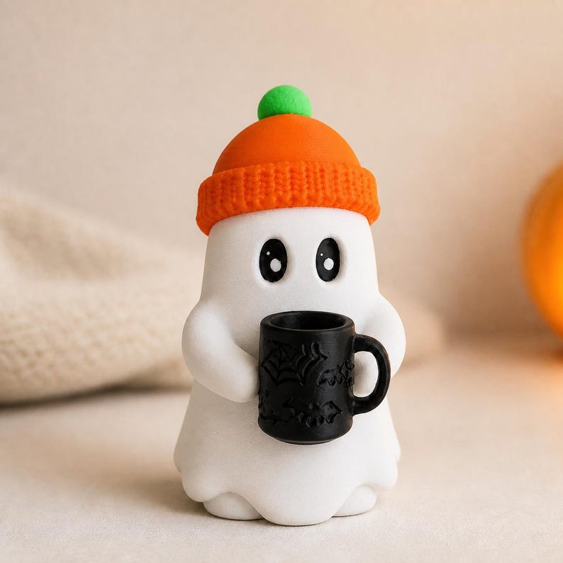 May include: A white ghost figurine wearing an orange knit beanie with a green pom-pom. The ghost has large black eyes and is holding a small black mug decorated with embossed spiderweb and bat patterns in its two rounded arms.