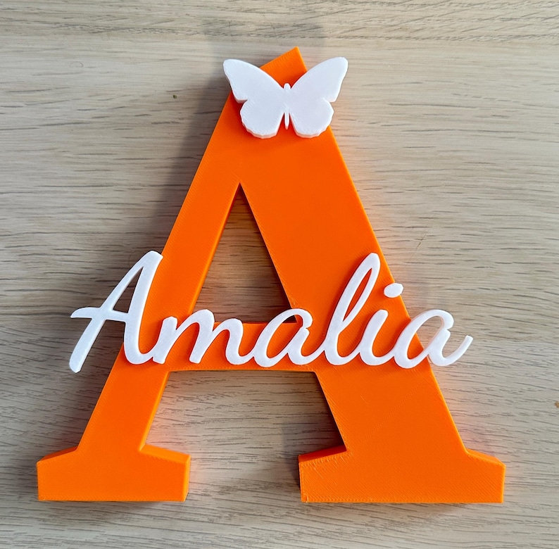 May include: A bright orange capital letter A featuring a white butterfly attached to the top center. The name Amalia is written in white cursive script across the front of the letter, creating a layered, three-dimensional decorative sign.