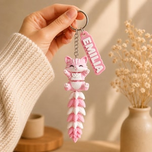 May include: A pink and white cat keychain featuring a segmented, flexible tail and a rectangular tag with the name EMILIA in white raised letters. The cat has a smiling expression, pointed ears, and a chain link attachment to a metal ring.