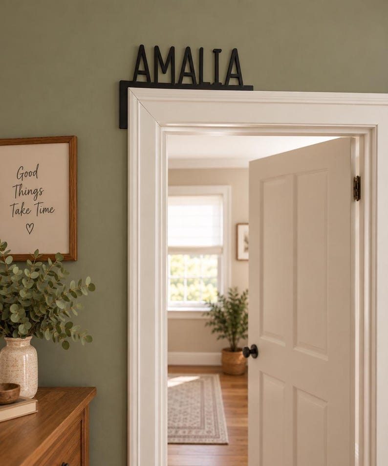 May include: A black L-shaped door corner sign featuring the name AMALIA in capital letters. The sign is mounted on the upper corner of a white door frame. The letters are sans-serif and have a flat, matte finish, positioned horizontally along the top.
