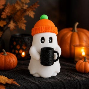 May include: A white ghost figurine wearing an orange knit beanie with a green pom-pom on top. The ghost has large black eyes and is holding a small black mug decorated with embossed spiderweb and bat patterns on its exterior surface.