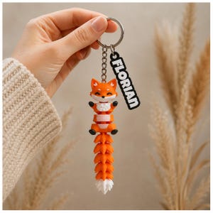 Könnte beinhalten: Ein flexibler Schlüsselanhänger in Form eines orange-weißen Fuchses mit segmentiertem Körper und Schwanz. Der Fuchs hat ein lächelndes Gesicht, schwarze Pfoten und eine weiße Brust. Ein silberner Ring trägt ein schwarzes Schild mit dem Text FLORIAN.