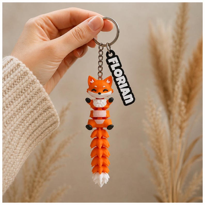 May include: A flexible orange and white fox keychain featuring a segmented body and tail. The fox has a smiling face, black paws, and a white chest. A silver metal ring attaches to a black rectangular tag with the white text FLORIAN printed on it.
