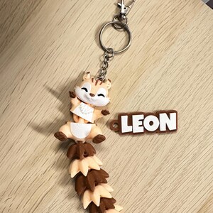 May include: A keychain featuring a small, articulated squirrel figure with a long, segmented tail in alternating light beige and brown colors. The squirrel has a white face and chest. Attached to the metal ring is a brown rectangular tag with the name LEON.