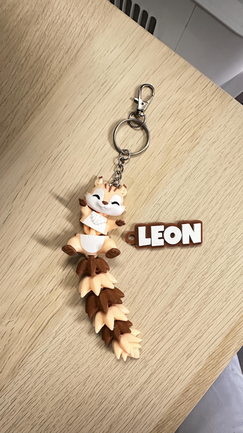 May include: A keychain featuring a small, articulated squirrel figure with a long, segmented tail in alternating light beige and brown colors. The squirrel has a white face and chest. Attached to the metal ring is a brown rectangular tag with the name LEON.