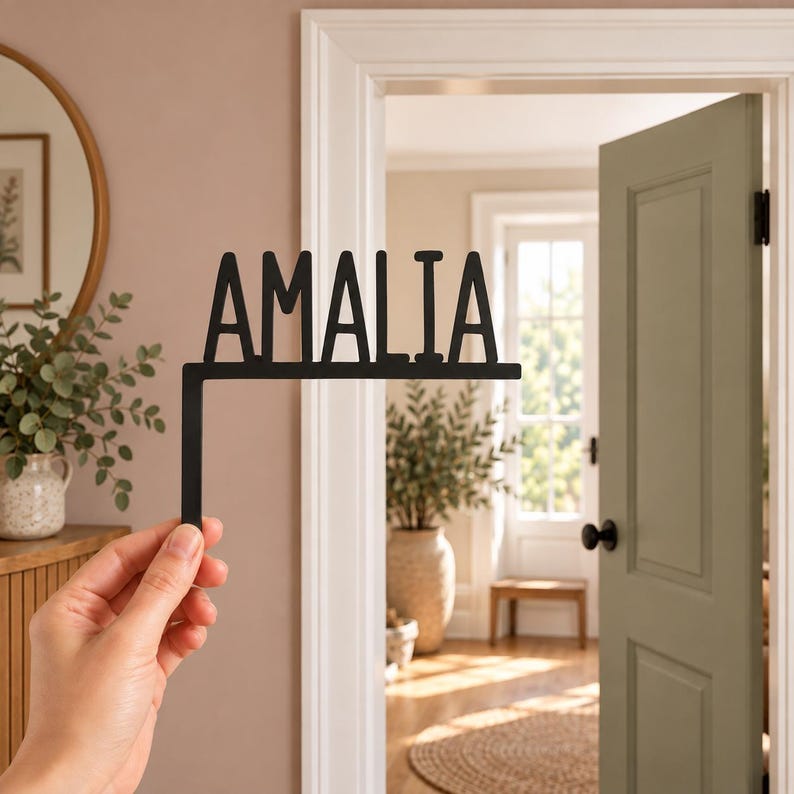 May include: A black metal door corner sign featuring the name AMALIA in a bold, sans-serif font. The sign has an L-shaped design intended to fit over the corner of a door frame, with the text extending horizontally from the vertical mount.