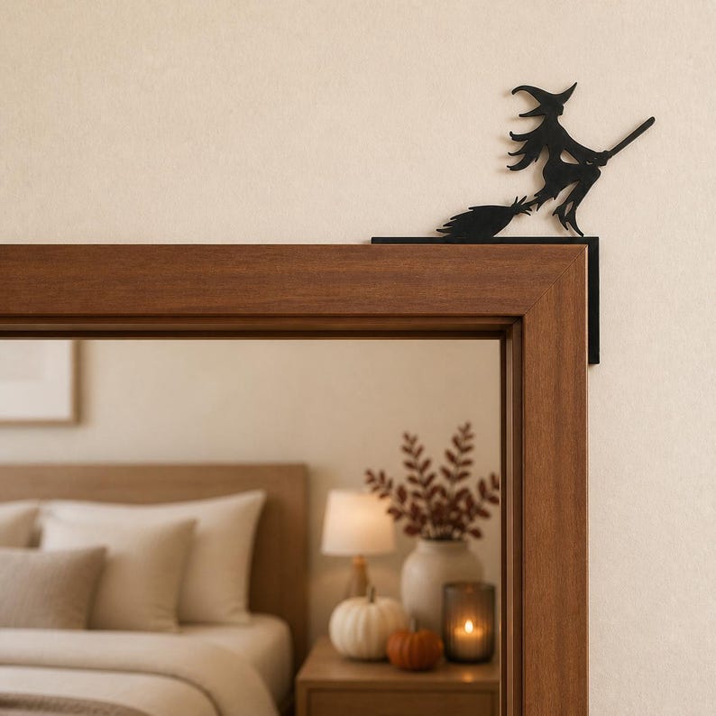 May include: A black metal silhouette of a witch riding a broomstick, designed to be mounted on the corner of a door frame. The flat, cutout figure depicts a pointed hat, flowing hair, and a broom, positioned to sit atop the wooden door frame edge.