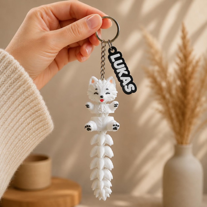 May include: A white fox keychain featuring a segmented, articulated tail and a small black tag with the name LUKAS in white capital letters. The fox has a smiling expression with a red tongue, black-tipped ears, and black paw pads on its feet.