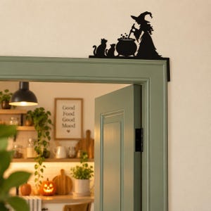 May include: A black metal door corner sign featuring a silhouette of a witch wearing a pointed hat, standing beside a bubbling cauldron and two cats. The decorative piece is designed to be mounted on the top corner of a door frame.