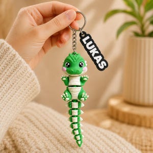 May include: A green and white articulated crocodile keychain featuring a small, cartoon-style reptile with large eyes and pink cheeks. Attached to the metal ring is a black rectangular tag with the white text LUKAS printed in bold letters.