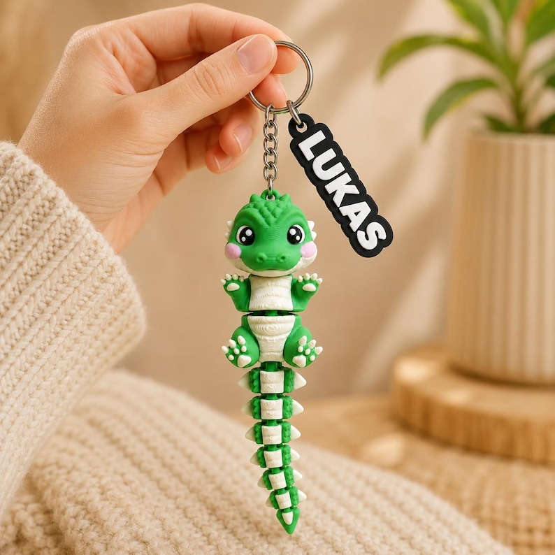 May include: A green and white articulated crocodile keychain featuring a small, cartoon-style reptile with large eyes and pink cheeks. Attached to the metal ring is a black rectangular tag with the white text LUKAS printed in bold letters.