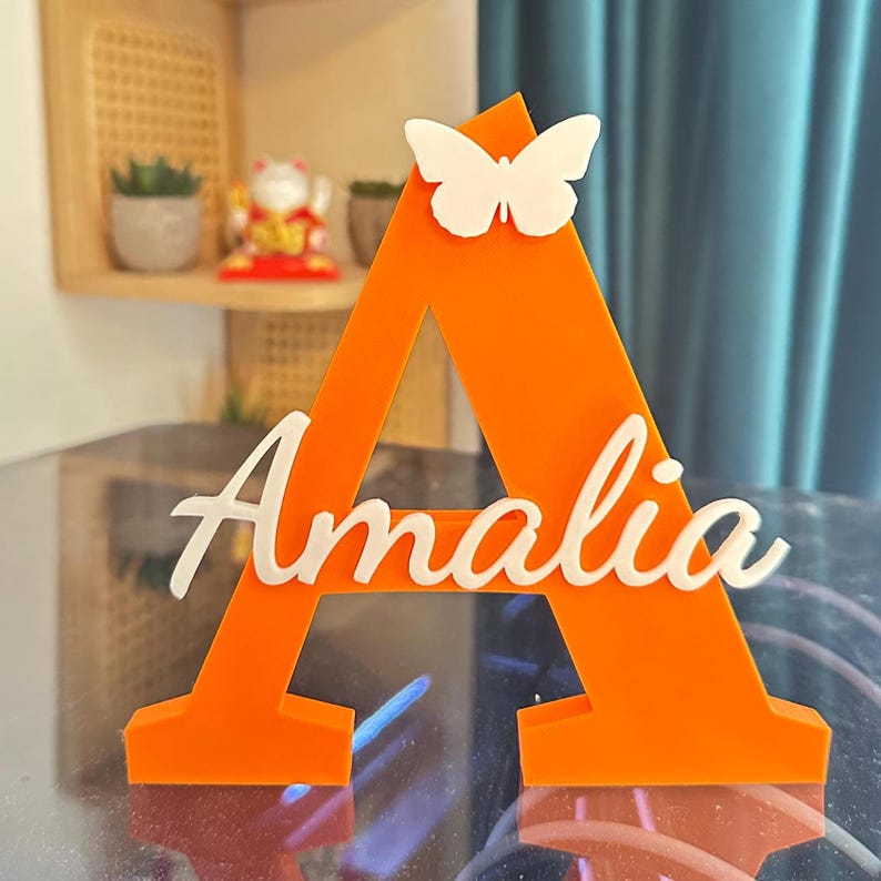 May include: A bright orange, three-dimensional letter A standing upright. The letter features a white butterfly attached to the top center and the name Amalia written in white cursive script across the middle section of the letter structure.