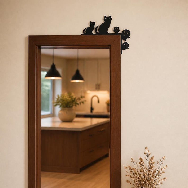 May include: A black metal decorative corner accent piece featuring silhouettes of a large cat, a small cat, and three ghosts. The item is mounted on the top right corner of a rectangular wooden door frame, contrasting against the light wall.