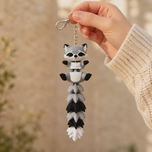 May include: A gray and white raccoon keychain featuring a multi-segmented, articulated tail that hangs vertically. The raccoon has black eye patches, small black paws, and a silver-toned metal lobster clasp attached to a short chain link connector.