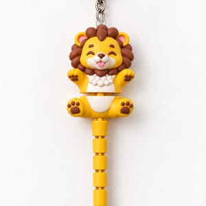May include: A keychain featuring a cartoon lion character with a yellow body, brown mane, and pink tongue. The lion is attached to a segmented, yellow, tail-like extension that ends in a brown tip. The entire figure hangs from a silver metal ring.