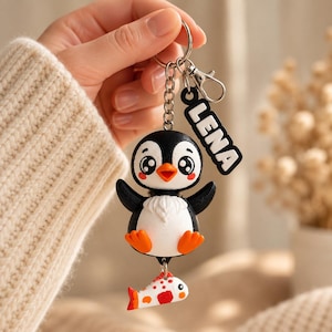 May include: A keychain featuring a black and white penguin character with orange feet and beak, attached to a silver metal ring and chain. A small black rectangular tag with the white text LENA is attached, along with a small white and orange fish charm.