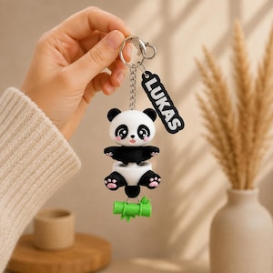 May include: A keychain featuring a black and white panda figure with pink paw pads, attached to a silver metal chain and ring. A black rectangular tag with the white text LUKAS is attached to the ring. A small green bamboo stalk hangs below.