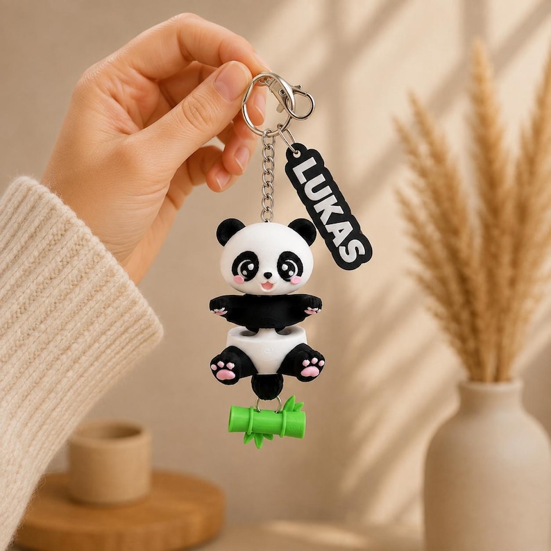 May include: A keychain featuring a black and white panda figure with pink paw pads, attached to a silver metal chain and ring. A black rectangular tag with the white text LUKAS is attached to the ring. A small green bamboo stalk hangs below.