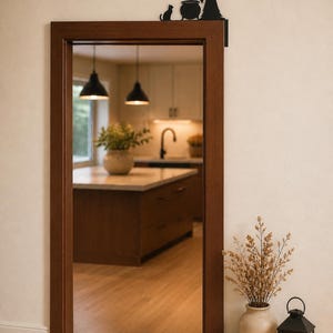 May include: A black metal door corner sign featuring a silhouette of a witch stirring a cauldron with a cat sitting beside it. The decorative piece is mounted on the upper right corner of a wooden door frame, showcasing a flat, cutout design.