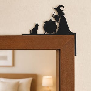 May include: A black metal door corner topper featuring a silhouette of a witch wearing a pointed hat, standing beside a cauldron and stirring it with a ladle, while a cat sits nearby on the flat base that rests along the top of the door frame.