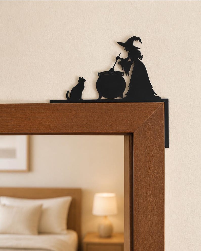 May include: A black metal door corner topper featuring a silhouette of a witch wearing a pointed hat, standing beside a cauldron and stirring it with a ladle, while a cat sits nearby on the flat base that rests along the top of the door frame.