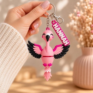 May include: A pink flamingo keychain featuring black and pink wings, a black beak, and a small rectangular tag with the name HANNAH printed in white letters. The flamingo is attached to a silver metal split ring and a lobster claw clasp.