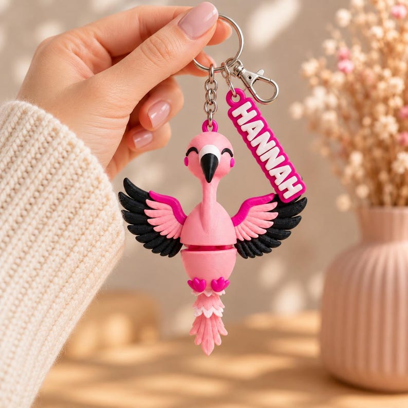 May include: A pink flamingo keychain featuring black and pink wings, a black beak, and a small rectangular tag with the name HANNAH printed in white letters. The flamingo is attached to a silver metal split ring and a lobster claw clasp.