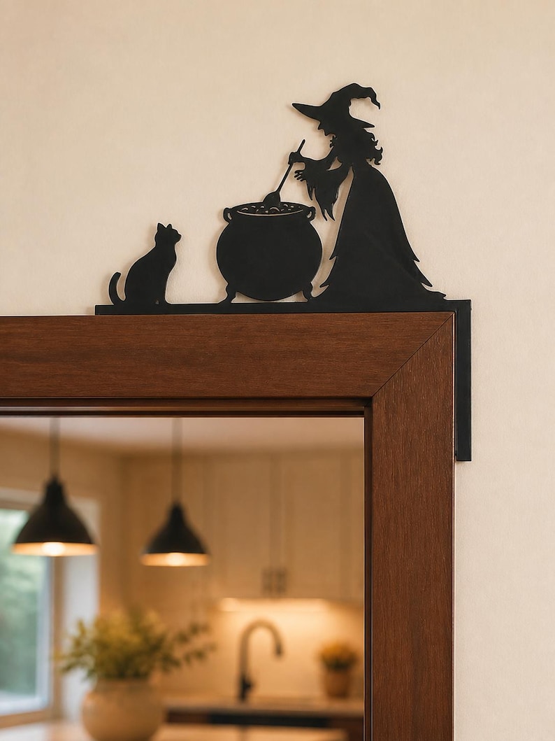 May include: A black metal door frame topper featuring a silhouette of a witch wearing a pointed hat, stirring a large cauldron with a spoon, and a cat sitting beside the pot. The decorative piece is designed to mount on the corner of a door frame.