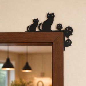 May include: A black metal door corner decoration featuring a silhouette of a large cat, a smaller cat, and three ghost figures. The flat, cutout design is mounted on the top right corner of a wooden door frame, creating a dark, graphic accent.