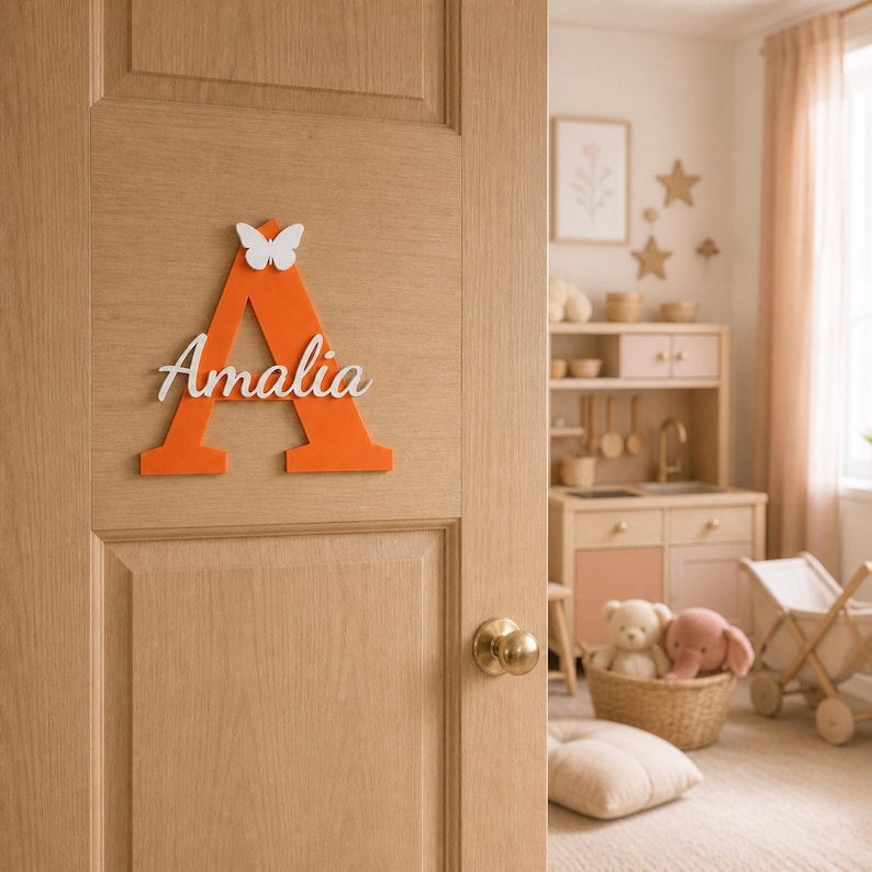 May include: A large orange wooden letter A mounted on a light wood door. The letter features a white butterfly cutout at the top and the name Amalia in white cursive script lettering across the center of the letter. The sign is flat and matte.