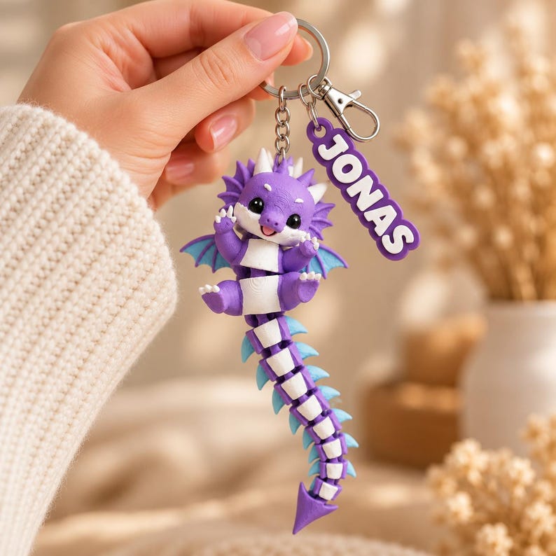 May include: A purple and white articulated dragon keychain featuring a long, segmented tail and a small dangling tag with the name JONAS in white letters on a purple background, attached to a silver metal keyring and lobster clasp.