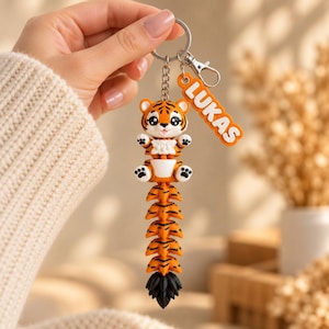 May include: A keychain featuring a cartoon tiger with orange and black stripes, a white belly, and a long articulated tail. Attached to the silver metal ring is a separate orange rectangular tag with the white embossed name LUKAS.