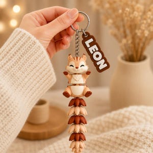 May include: A keychain featuring a light brown and cream-colored squirrel figure with a long, articulated, segmented tail in alternating shades of brown and cream. A brown rectangular tag with the white text LEON is attached to the metal ring.