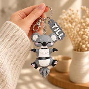 May include: A gray and white koala keychain featuring a split-body design, a black nose, pink cheeks, and black paws with pink pads. A gray rectangular tag attached to the silver metal ring displays the white text TILL in capital letters.