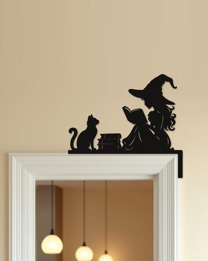May include: A black metal wall art piece designed to sit on a door frame corner, featuring a silhouette of a witch wearing a pointed hat and reading an open book, accompanied by a cat sitting next to a small stack of books on a flat base.