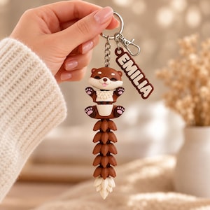 May include: A brown and white articulated otter keychain featuring a long, segmented tail and a small dangling tag with the name EMILIA printed in white letters. The otter has a smiling face, raised paws with pink pads, and a metal split ring attachment.