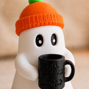 May include: A white ghost figurine with large black eyes wearing an orange beanie with a green pom-pom. The ghost is holding a small black mug decorated with embossed patterns of spiderwebs, bats, and pumpkins on its exterior surface.